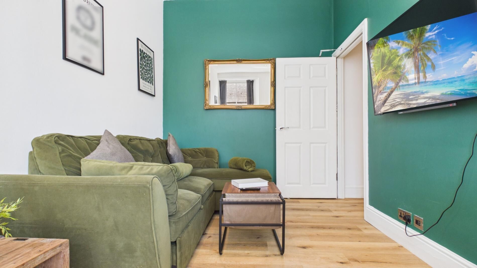 Un salon avec un canapé vert, une petite table, un tableau encadré, une porte blanche et un téléviseur fixé au mur qui diffuse une scène de plage.
