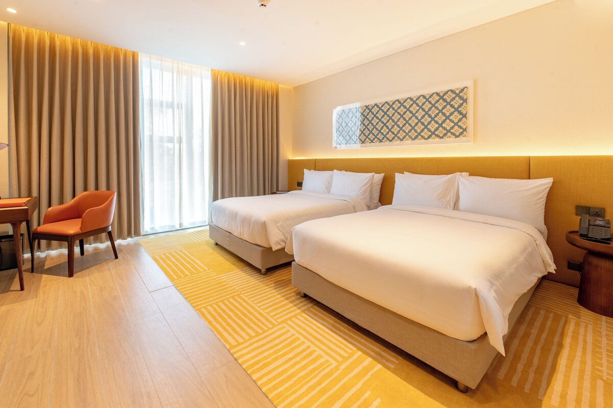 A hotel room with two beds, a desk, a chair, and a large window with curtains.