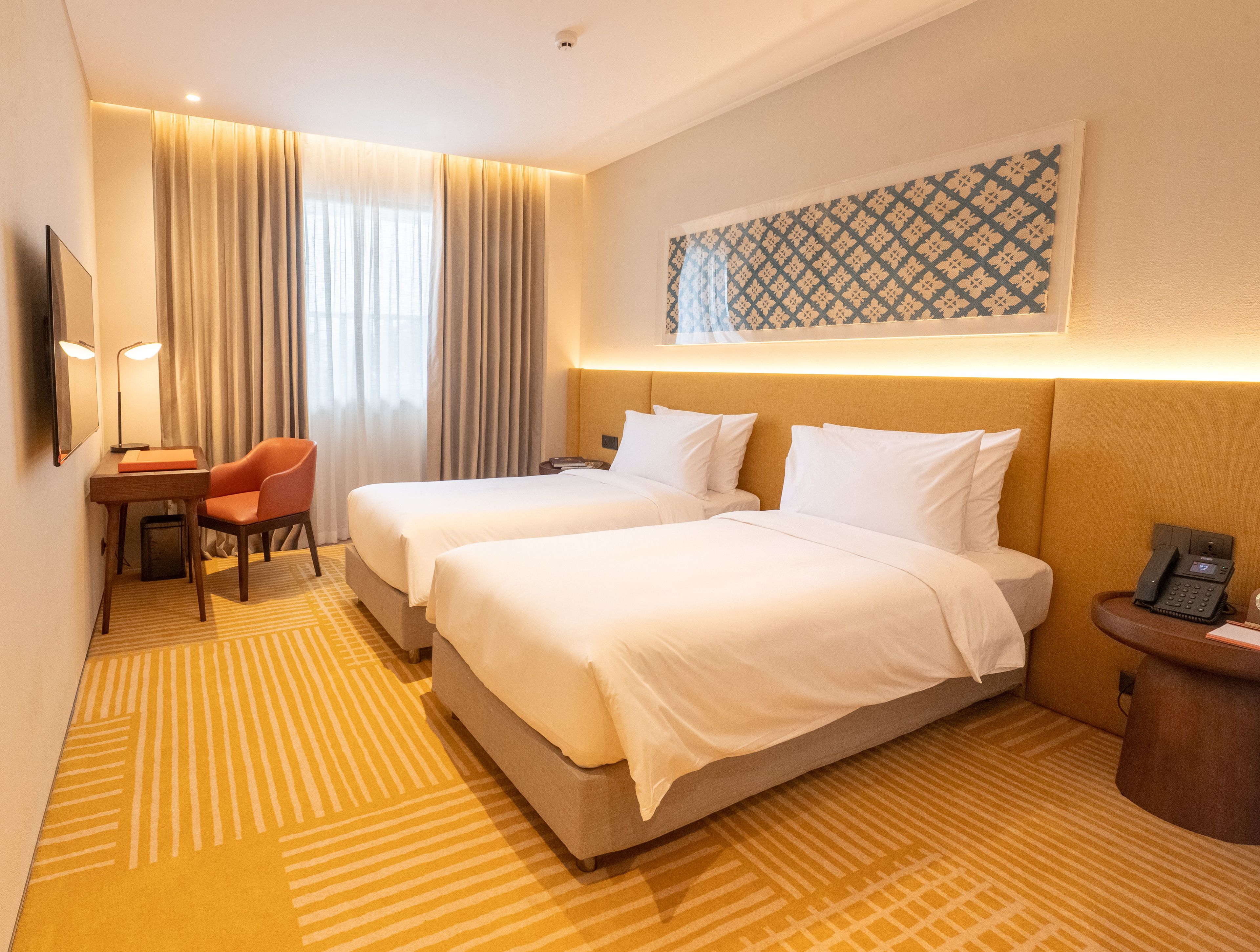 A hotel room with two beds, a desk with a chair, a telephone, and a wall-mounted artwork.