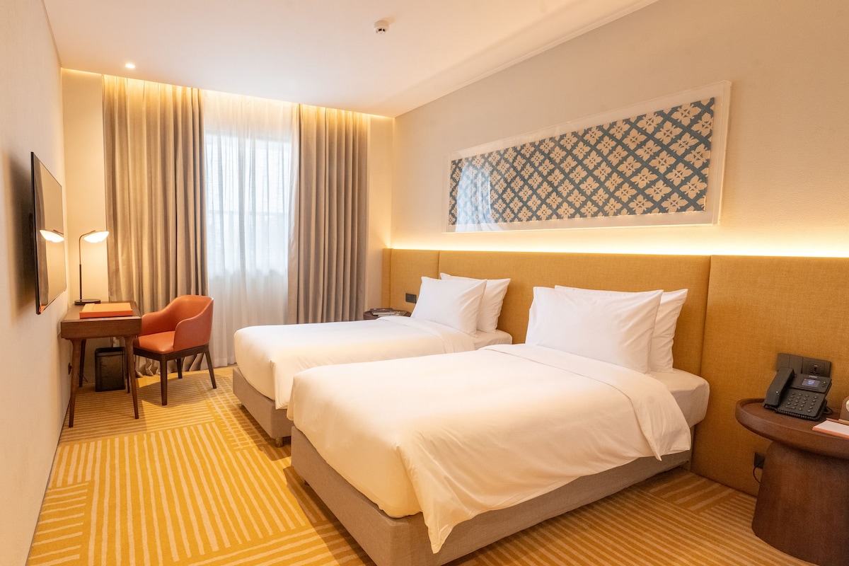 A hotel room with two beds, a desk with a chair, a telephone, and a wall-mounted artwork.