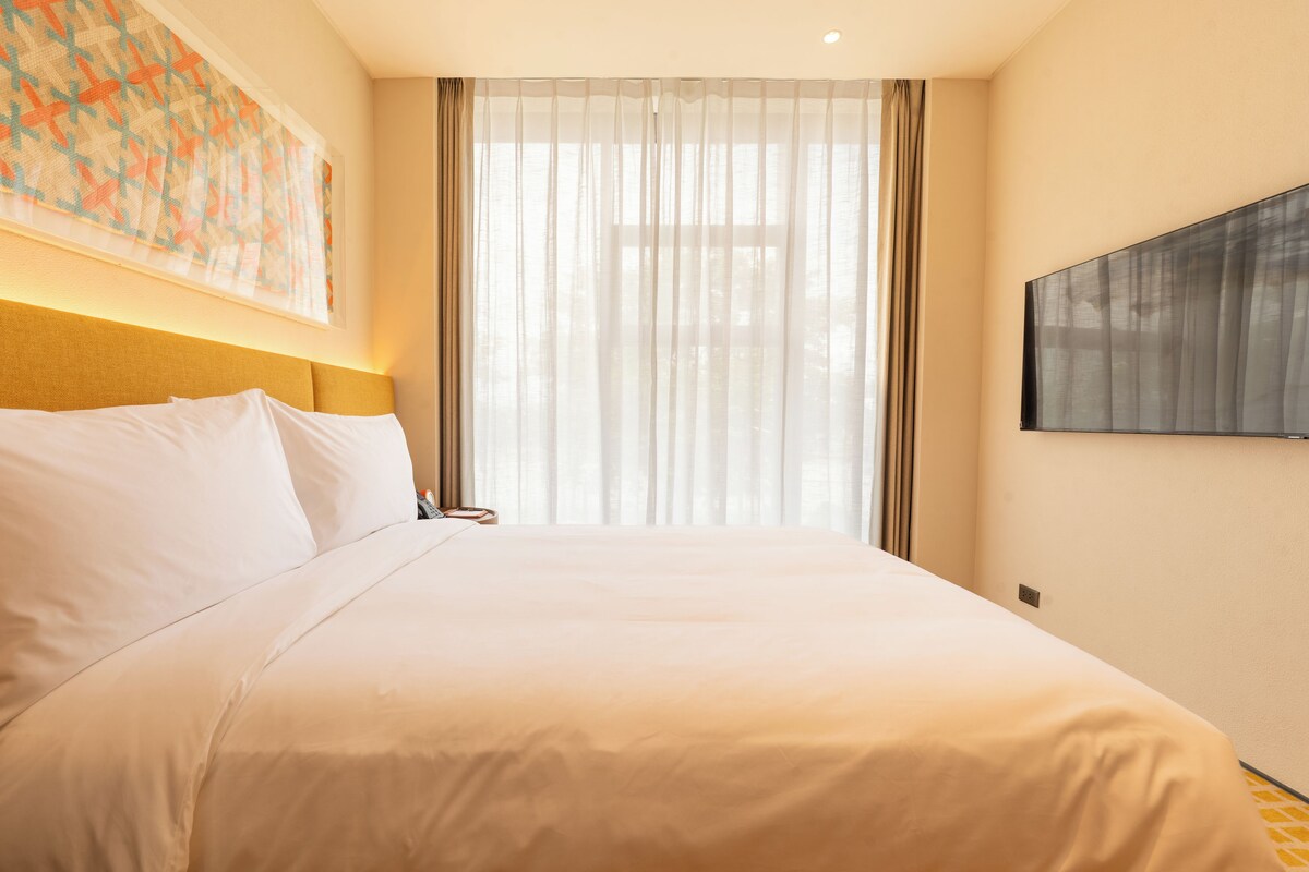 A hotel room with a large bed, a mounted TV, and a window with sheer curtains.