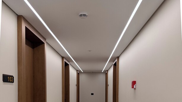 Un couloir avec des portes en bois, un détecteur d’incendie et un panneau indiquant le numéro des pièces.