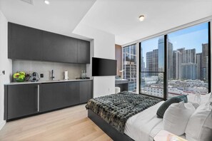 Ein modernes Schlafzimmer mit einem großen Bett, Blick auf die Stadt, einer Küche mit dunklen Schränken und einem Flachbildfernseher.