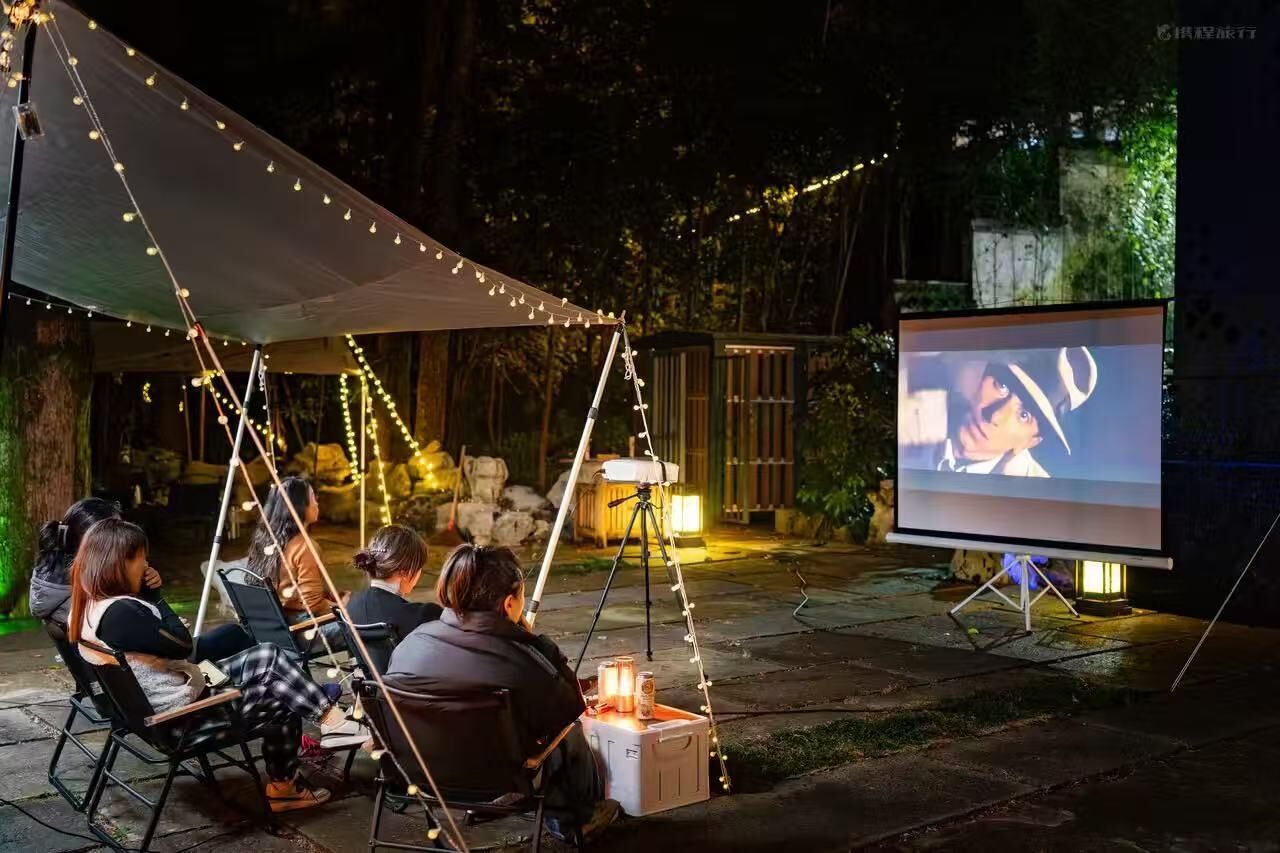 An outdoor movie screening setup with a projector and screen, surrounded by string lights and seating.