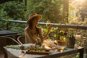 Uma pessoa sentada em uma mesa ao ar livre, com diversos pratos e bebidas, em meio à vegetação.