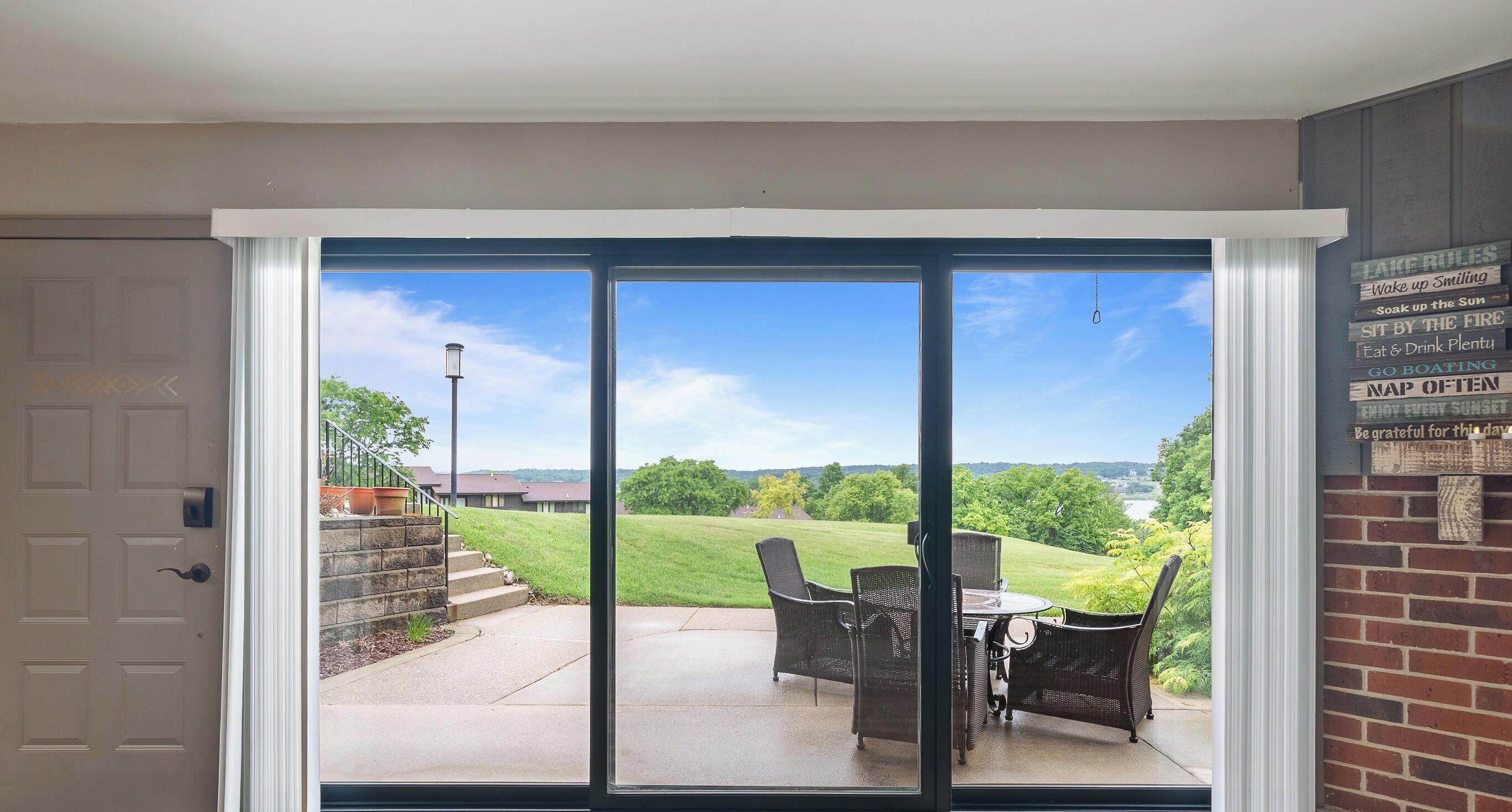 A sliding glass door leading to an outdoor patio with a table and chairs, a brick wall, and a view of a green landscape.