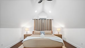 Un dormitorio con cama, dos mesitas de noche, ventilador de techo y ventana con cortinas.