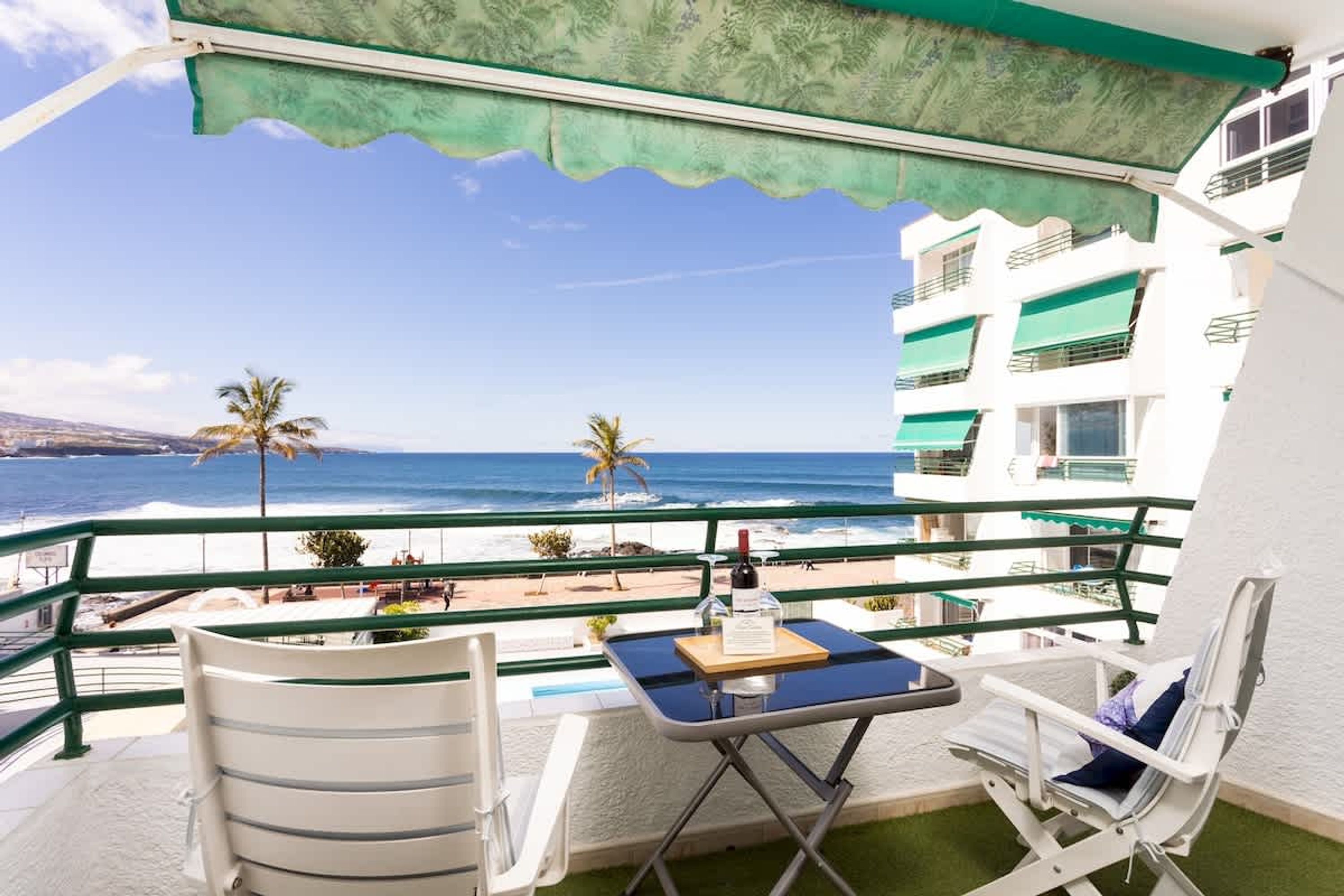 A balcony with a view of the beach and ocean, a table with a bottle and menu, and two chairs.