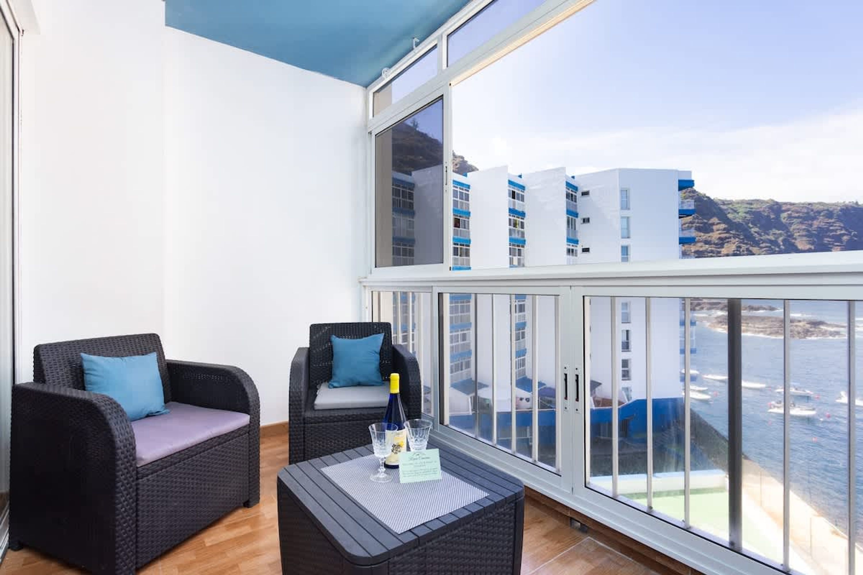 A balcony with wicker furniture, a glass table with a bottle and glasses, and a view of buildings and water.
