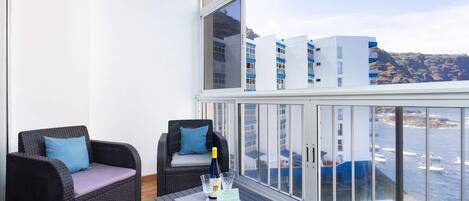 A balcony with wicker furniture, a glass table with a bottle and glasses, and a view of buildings and water.