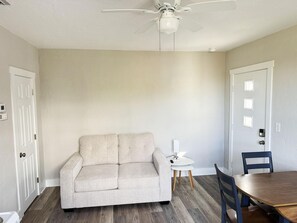 Un salon avec un canapé beige, une table à manger avec des chaises, un ventilateur de plafond et une petite table d’appoint blanche.
