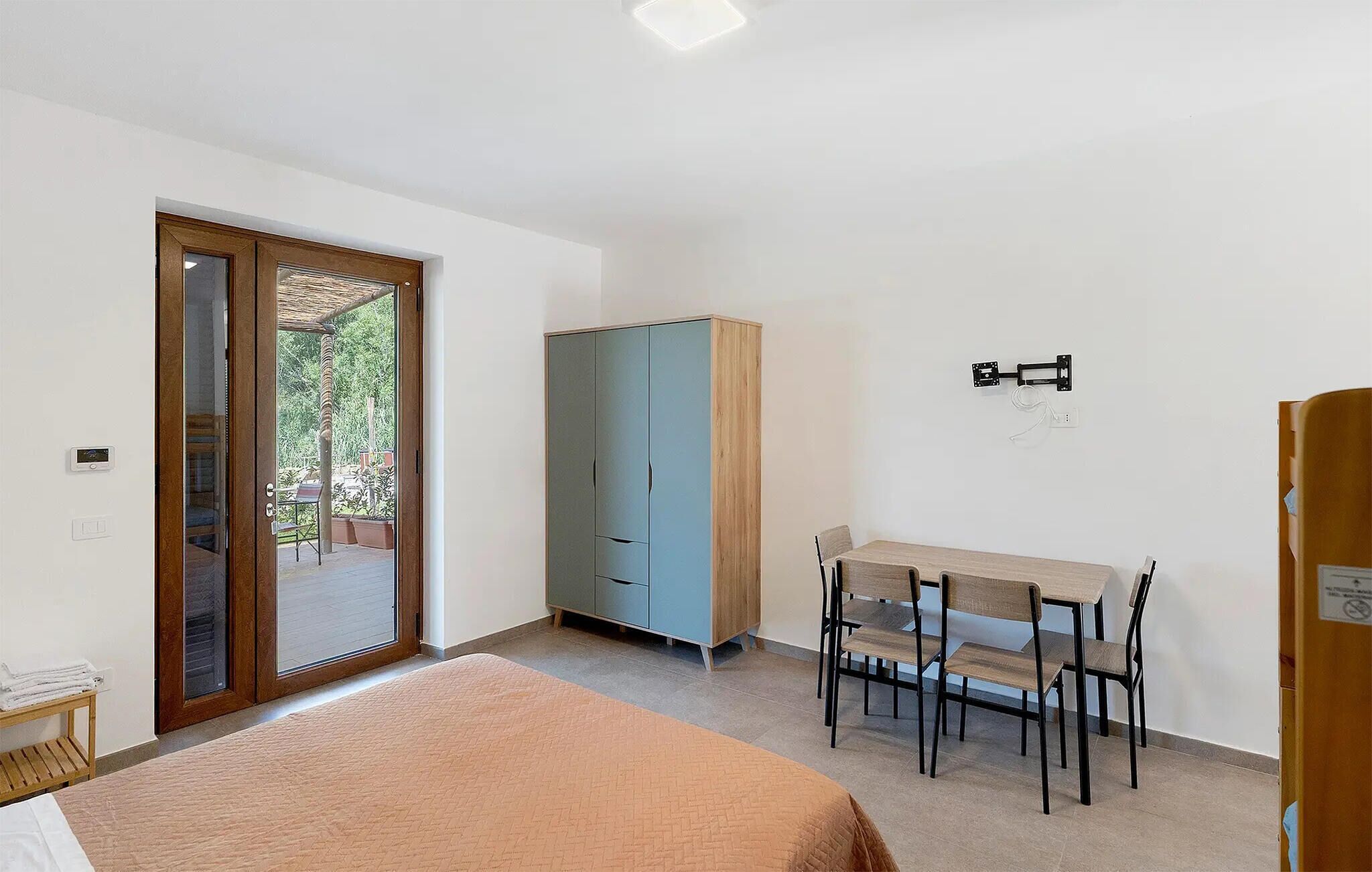 Un dormitorio con cama, armario, escritorio con sillas y puerta corrediza de vidrio que da a un espacio exterior.
