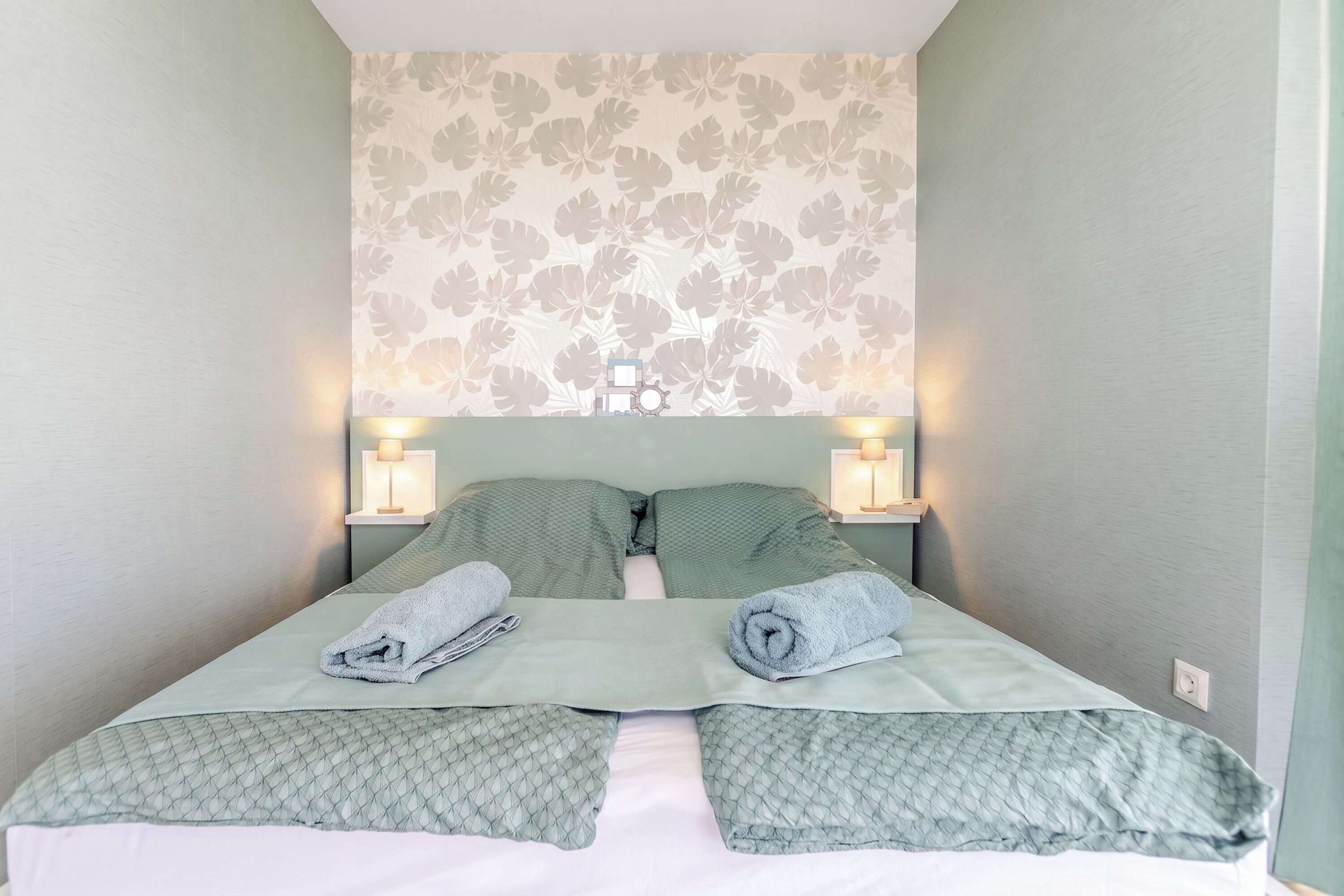 Una cama bien hecha con dos almohadas y dos toallas enrolladas, flanqueada por dos lámparas de noche sobre una pared con estampado floral.