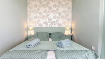 Una cama bien hecha con dos almohadas y dos toallas enrolladas, flanqueada por dos lámparas de noche sobre una pared con estampado floral.