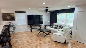 A living room with a white sofa, a TV, a ceiling fan, and a dining area with chairs and a table.