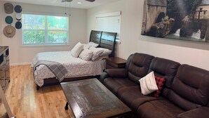 A bedroom with a bed, a ceiling fan, a window, a painting, a brown leather sofa, and a wooden coffee table.