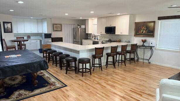 A modern kitchen with white cabinets, a large island with bar stools, a dining area with a table and chairs, and a living room with a sofa.