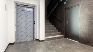 A modern interior with a grey tiled floor, a staircase with a dark wall, and a door with directional signs. - Mostek Riverside Deluxe Studio (Gdańsk)
