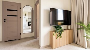 A modern living room with a flat-screen TV mounted on a wooden console, a door, and a mirror reflecting a kitchen area. - Rumia Stylish Apartment (Rumia)