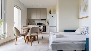 A modern hotel room with a bed, a dining area with chairs, a kitchen with appliances, and a large window. - Rumia Stylish Apartment (Rumia)