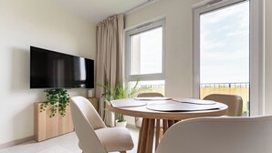 A modern living room with a flat-screen TV mounted on the wall, a round wooden dining table with chairs, a potted plant, and a view of the outside through large windows. - Rumia Stylish Apartment (Rumia)