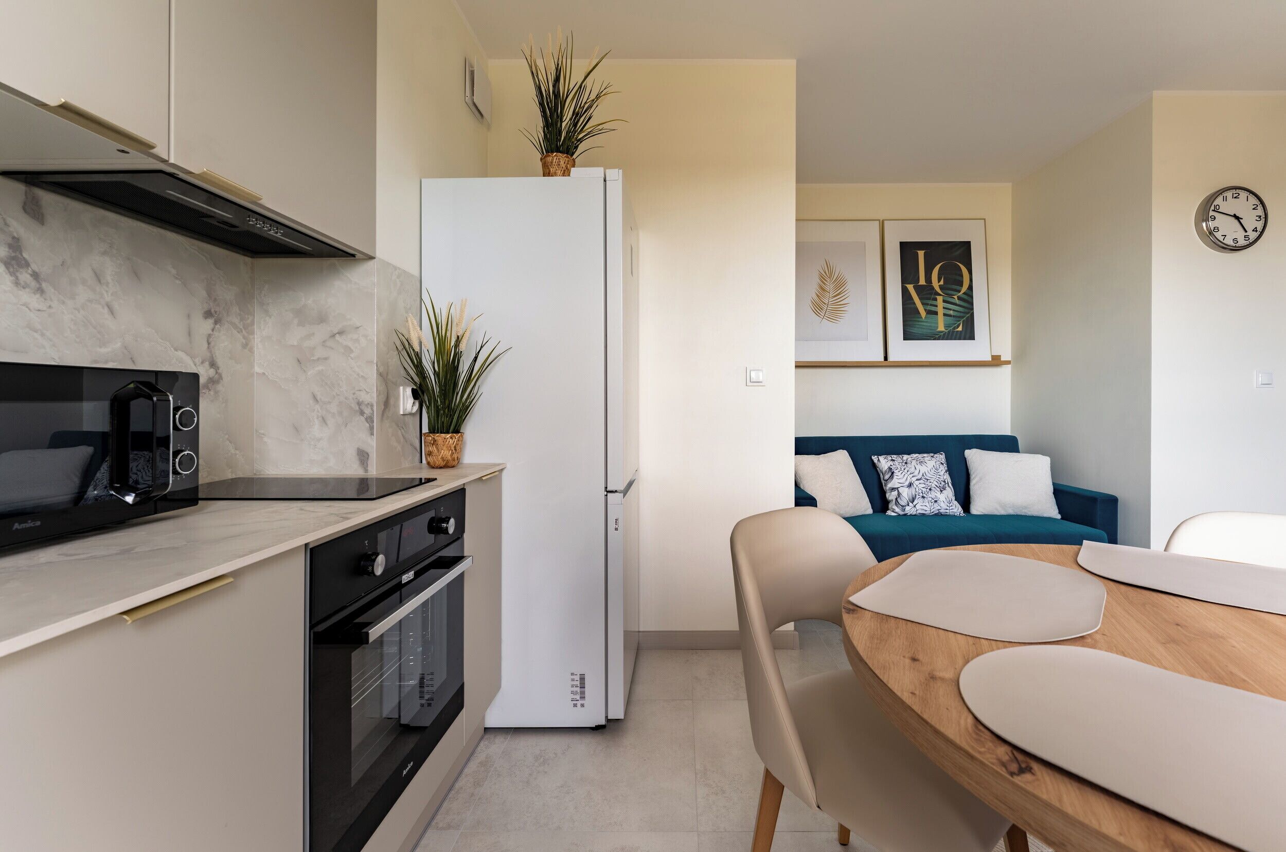 A modern kitchen with a built-in oven, microwave, and white refrigerator. There is a dining table with chairs and a blue sofa in the background.