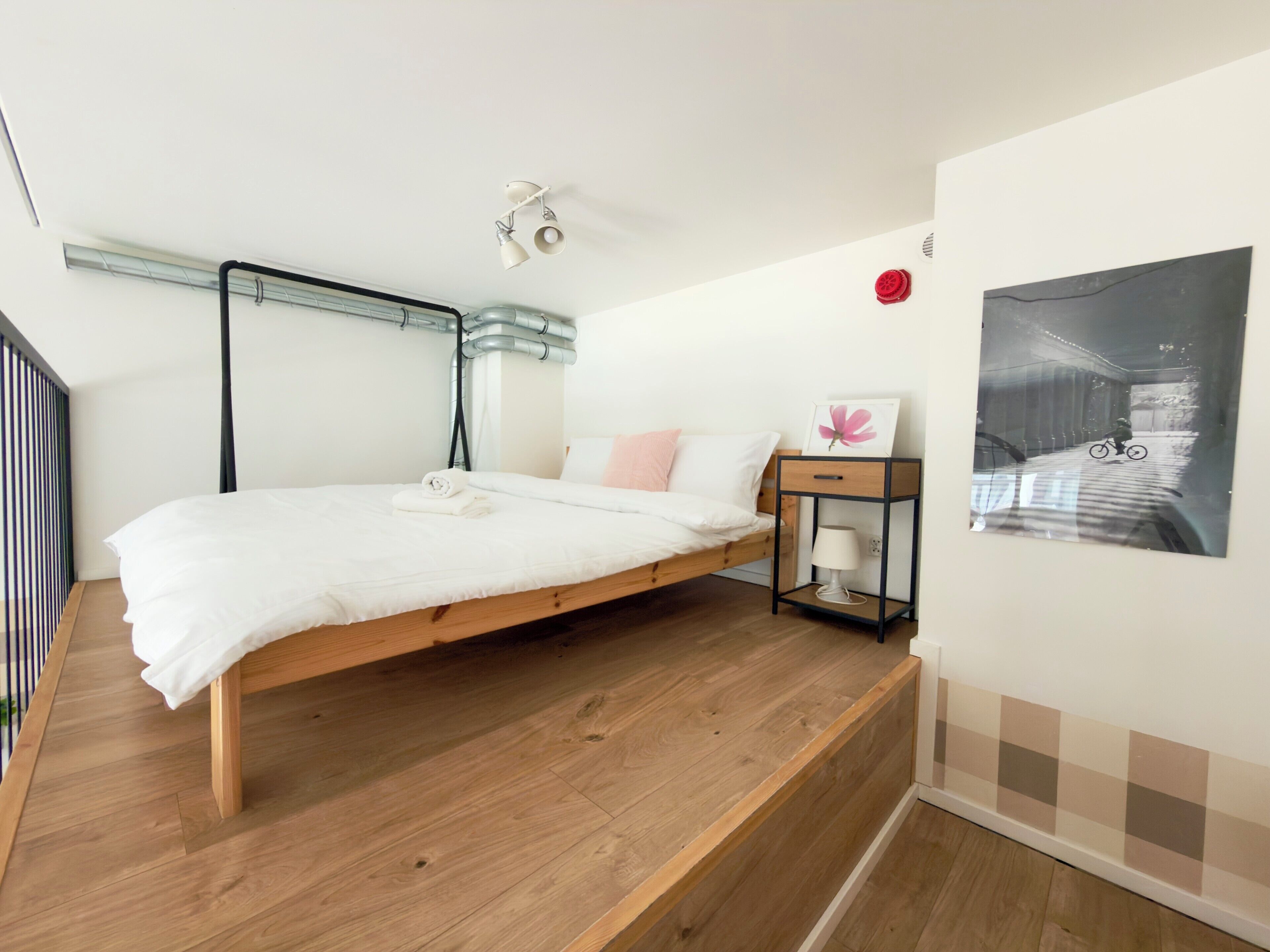 Un dormitorio con cama de madera, una mesita de noche con lámpara, un cuadro enmarcado en la pared y una ventana con persianas.