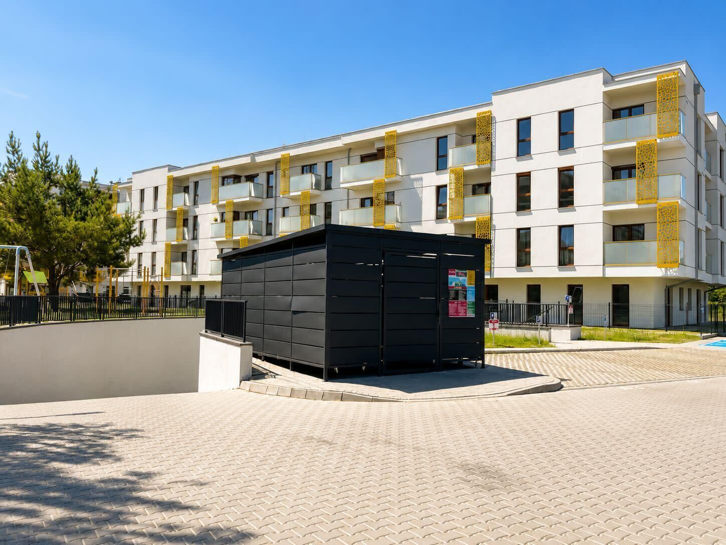 A modern apartment building with balconies, a small black structure, and a paved area.
