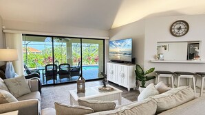 A living room with a sofa, coffee table, TV, and a view of a pool and greenery. - Golf Retreat - Pool, Spa & Fire Pit (Rotonda West)