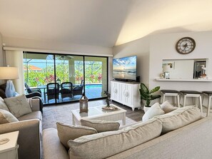 A living room with a sofa, coffee table, TV, and a view of a pool and greenery. - Golf Retreat - Pool, Spa & Fire Pit (Rotonda West)