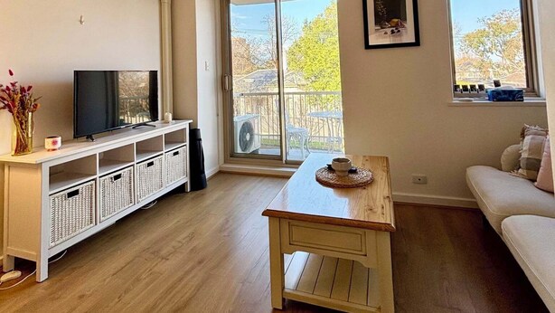 Ein Wohnzimmer mit Fernseher, weißen Möbeln, einem Holztisch und einer Glasschiebetür, die zu einem Außenbereich führt.