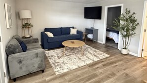 Una sala de estar con un sofá azul, un sofá gris, una mesa de centro, un televisor, una planta y una puerta que da a un dormitorio.