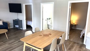 Una sala moderna con una mesa de comedor de madera, sillas blancas, un televisor de pantalla plana, una pequeña mesita auxiliar y un sofá.