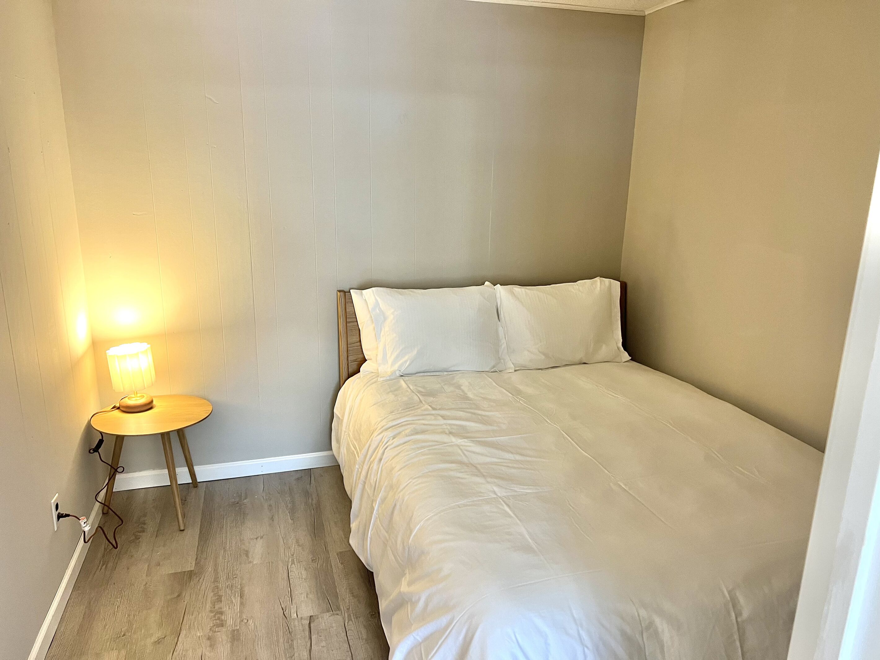 A small, neatly made bed with white bedding in a room with a wooden floor and a wall-mounted lamp.