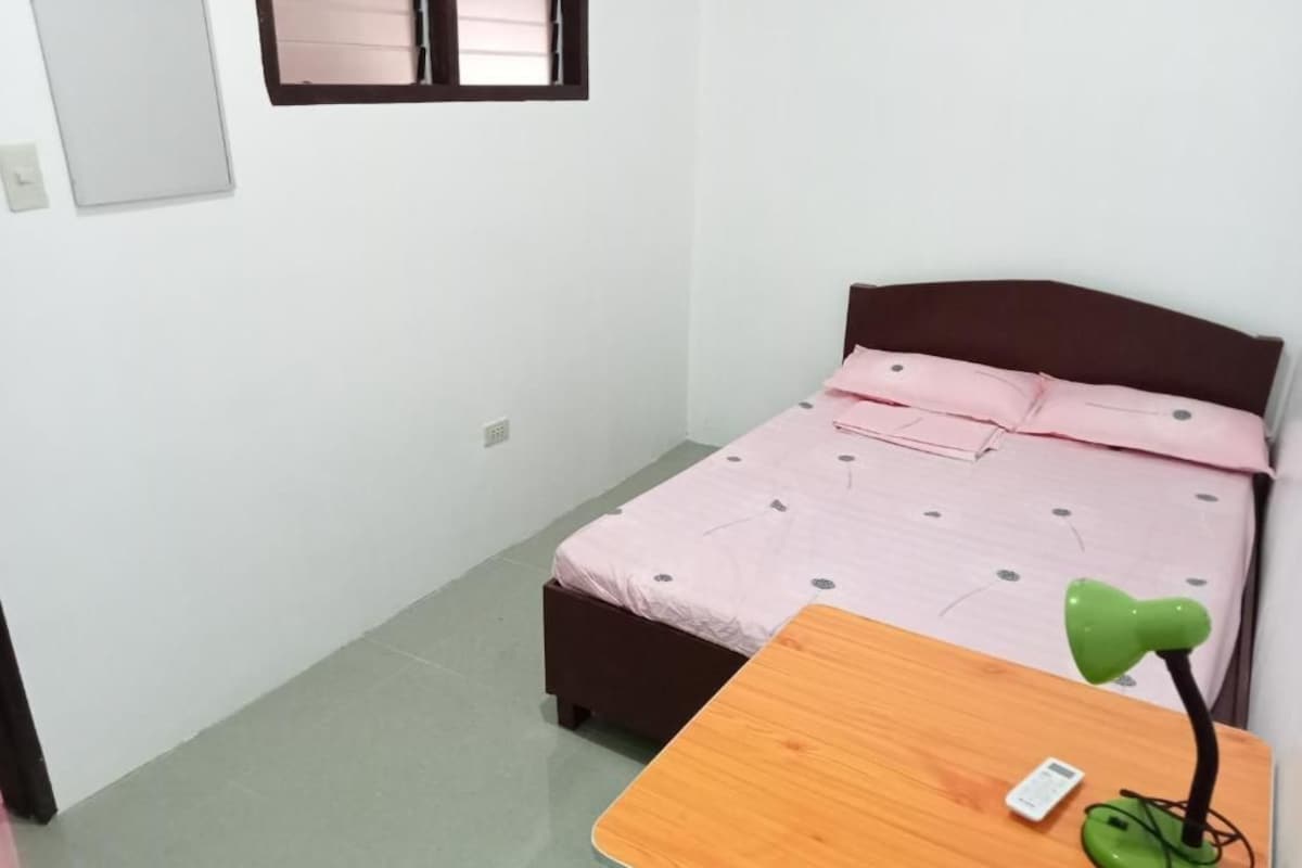 A bedroom with a bed, a wooden table, a green desk lamp, and a window.