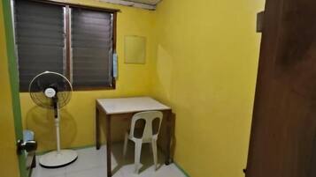 A small room with yellow walls, a white table, a white chair, a standing fan, and a window with a closed shutter.