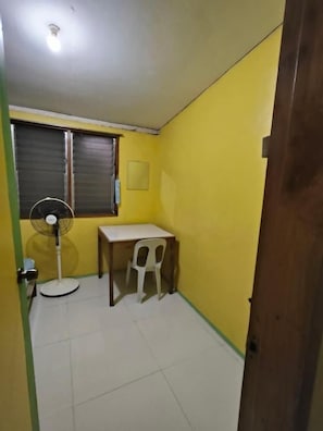 A small room with yellow walls, a white table, a white chair, a standing fan, and a window with a closed shutter. - RGR CAMIGUIN TRAVEL AND PENSION HOUSE (Mambajao)