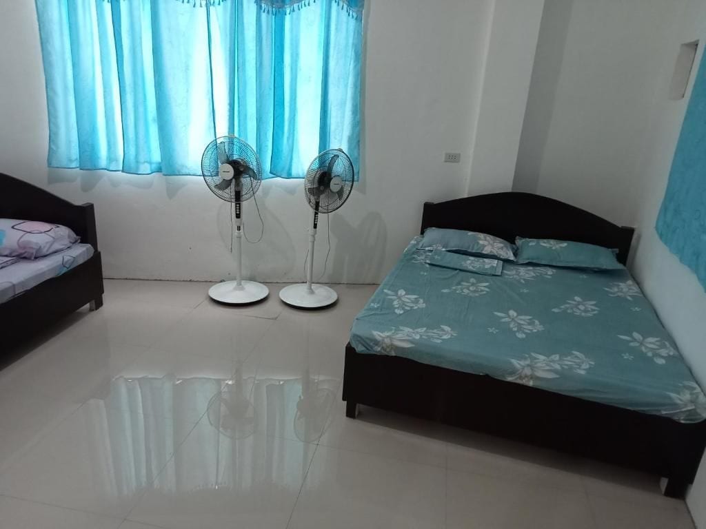 Quarto com dois ventiladores, cama com colcha florida e janela com cortinas azuis.