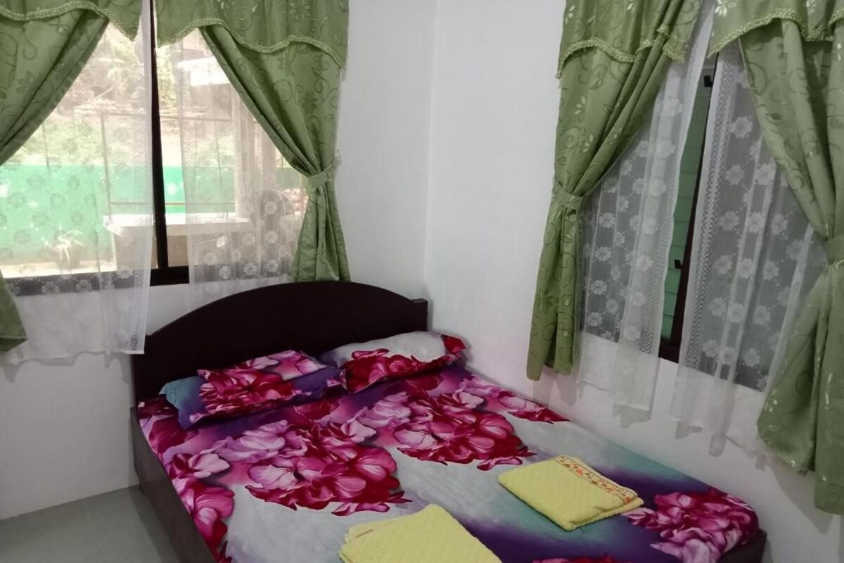 A single bed with a floral bedspread, green curtains, and a window with a view of greenery outside.