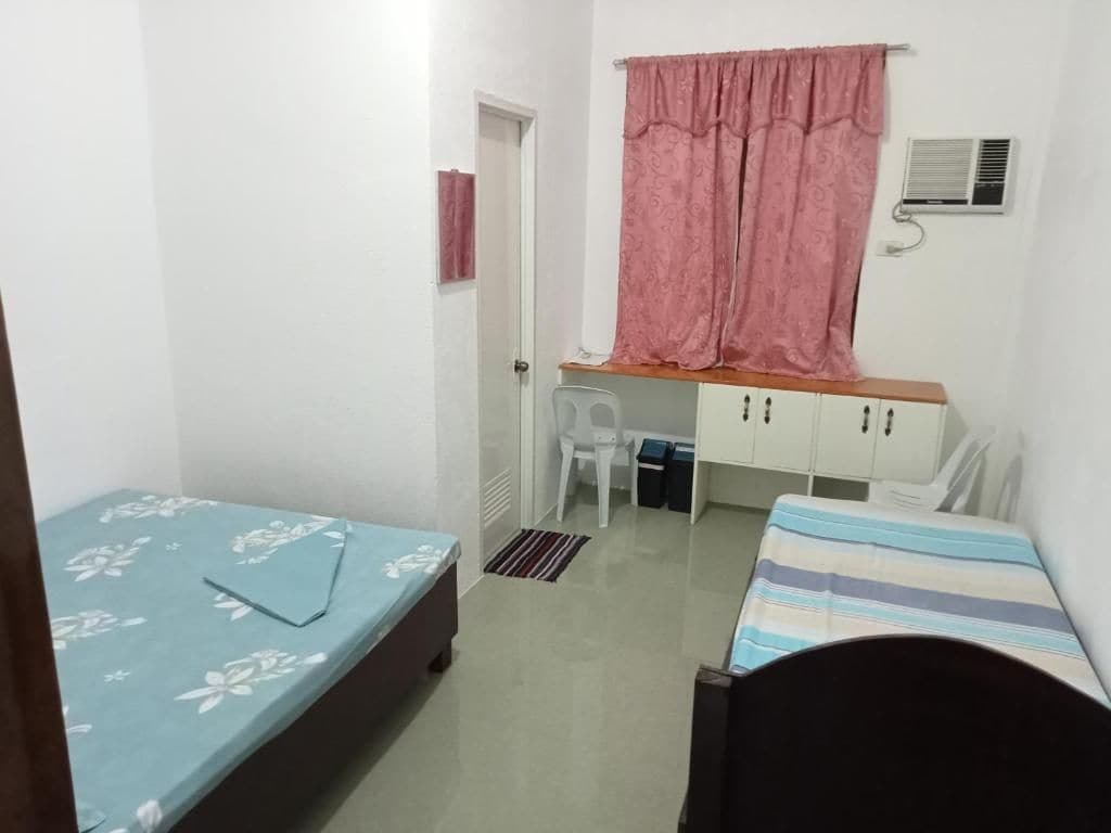 A small, compact room with a bed, a desk, and a chair. There is a window with a curtain, an air conditioning unit, and a door.