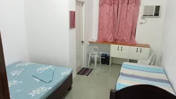 Una habitación pequeña y funcional con cama, escritorio y silla. Tiene ventana con cortina, unidad de aire acondicionado y puerta.