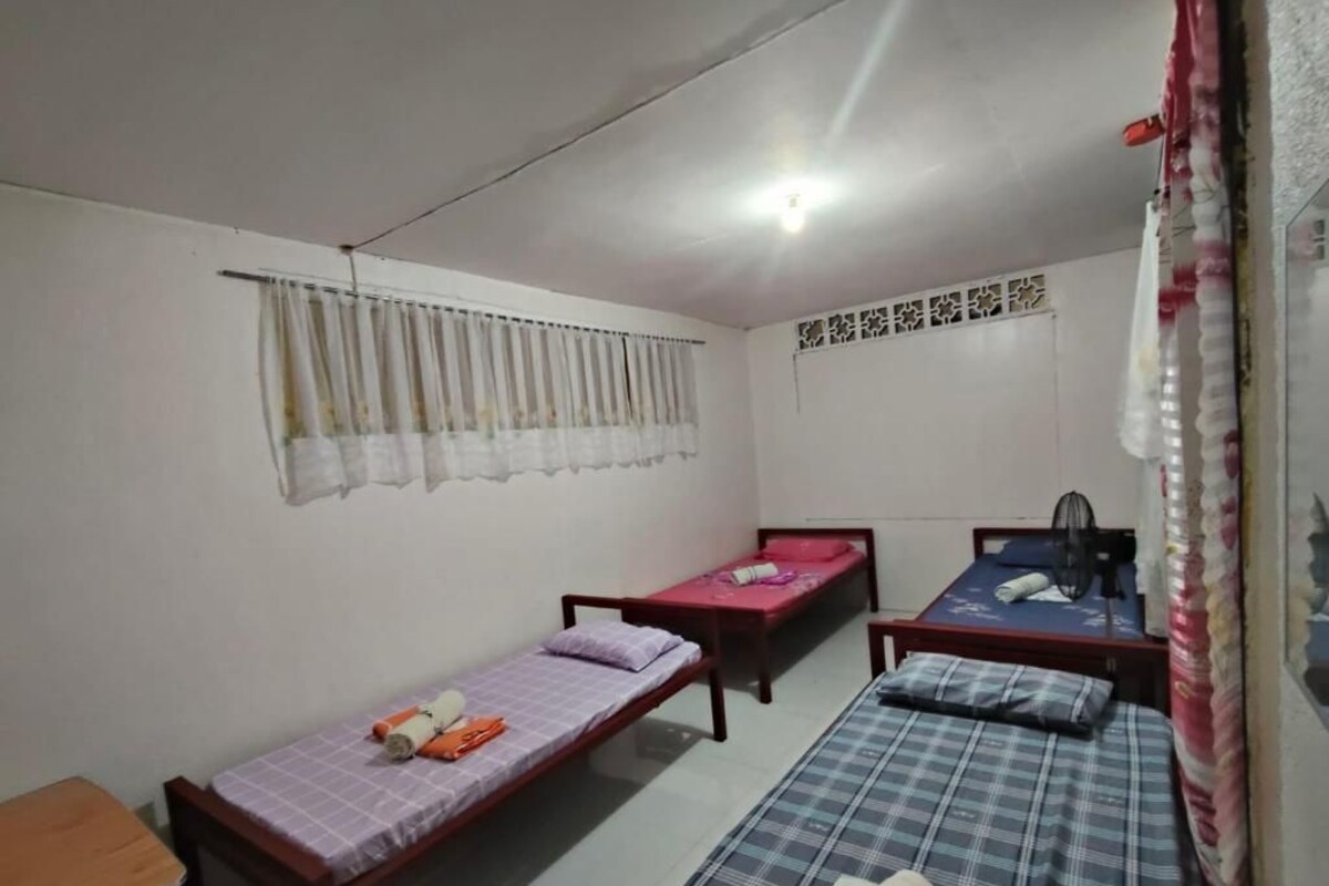 A dormitory room with three single beds, a wooden bench, a ceiling fan, and a curtain.