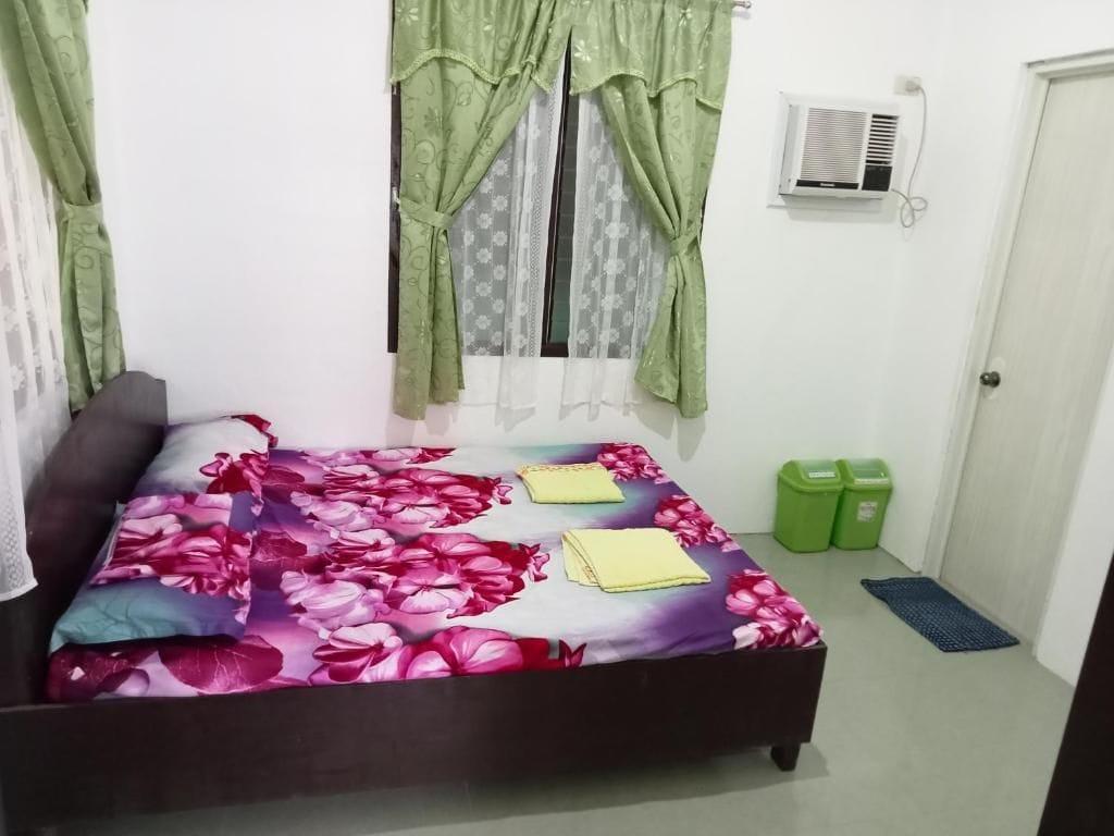 A bedroom with a bed, curtains, an air conditioner, a door, and a trash bin.