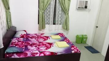 A bedroom with a bed, curtains, an air conditioner, a door, and a trash bin.