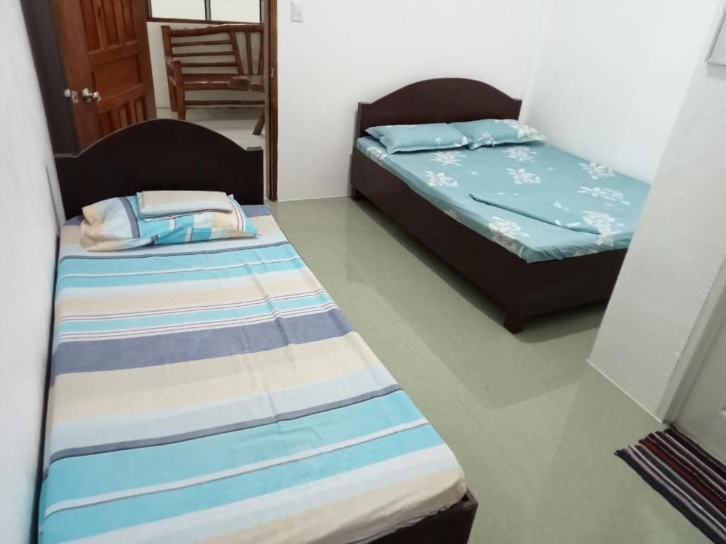 A small room with two single beds, one with a striped bedspread and the other with a floral bedspread. There is a wooden chair and a door visible.