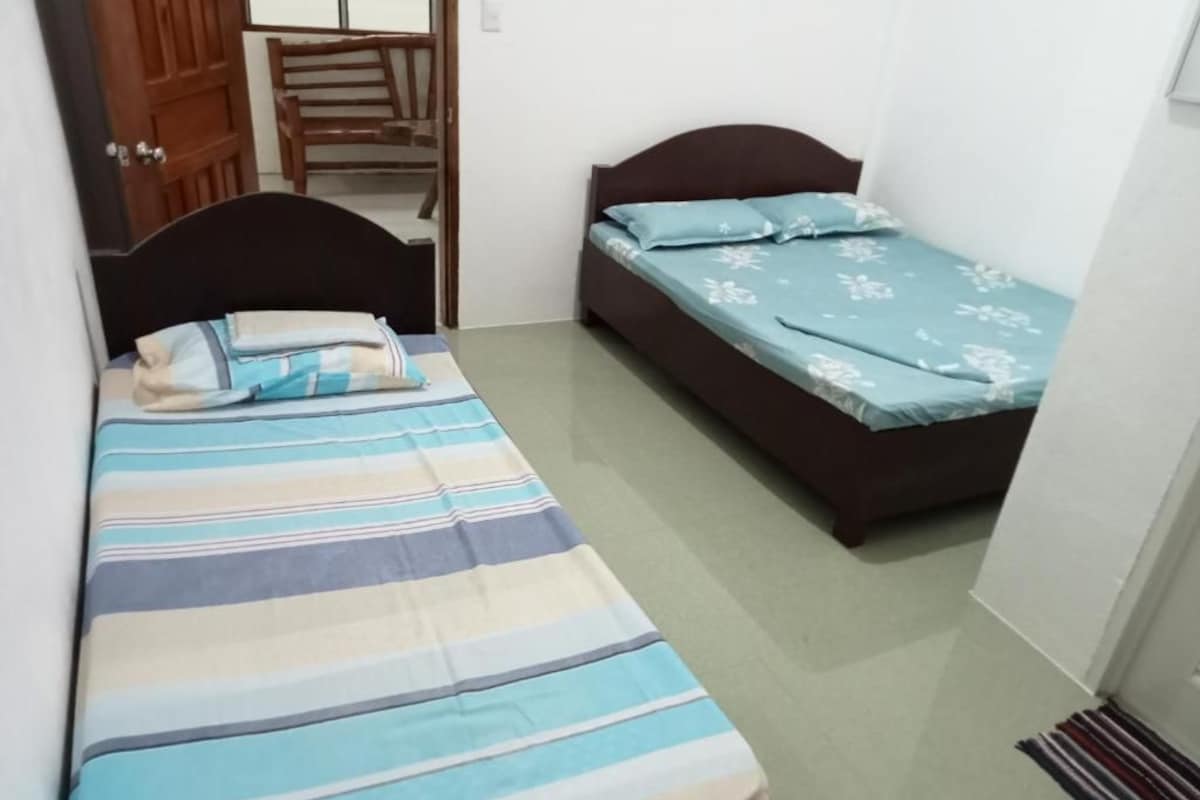 A small room with two single beds, one with a striped bedspread and the other with a floral bedspread. There is a wooden chair and a door visible.