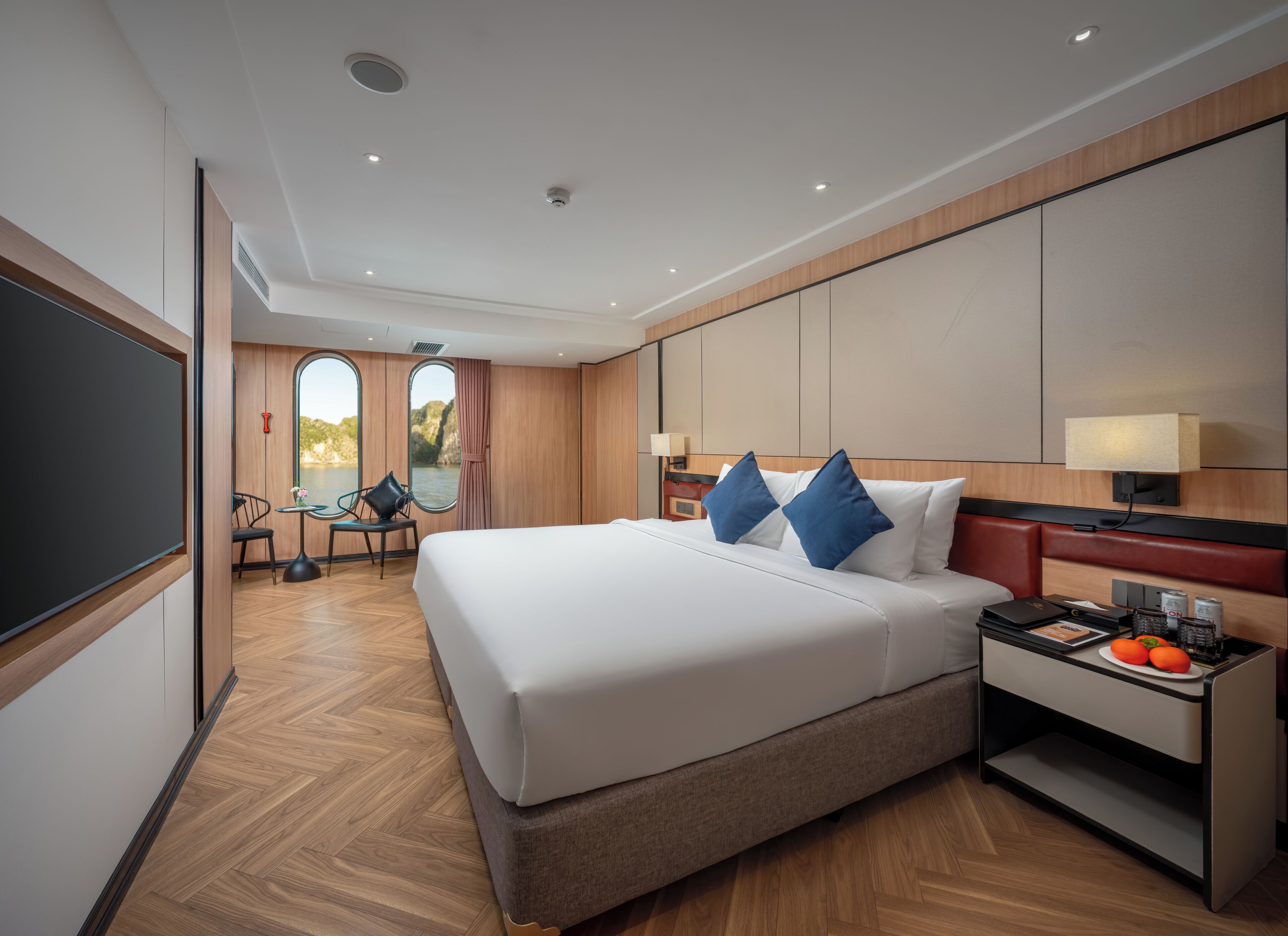 A modern hotel room with a large bed, a desk with a lamp, a chair, and a view of a body of water through a window.