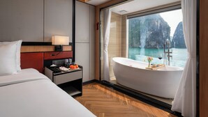 A hotel room with a large bed, a desk with a lamp, a minibar, and a bathtub with a view of the sea and cliffs.