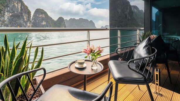 A deck with a view of the sea and mountains, a table with a cup and flowers, and a chair.