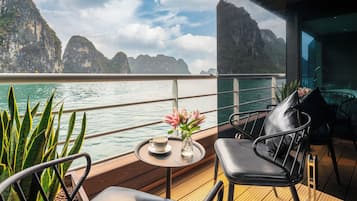 A deck with a view of the sea and mountains, a table with a cup and flowers, and a chair.